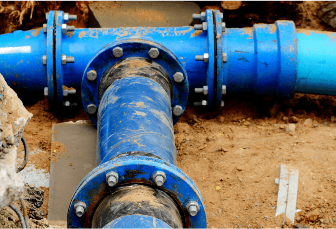Close-up view of a blue trunk water main pipe junction partially buried in soil, illustrating critical infrastructure at risk of leakage, relevant to Ofwat's updated regulatory focus on trunk main leakage detection, monitoring, and repair innovations. 